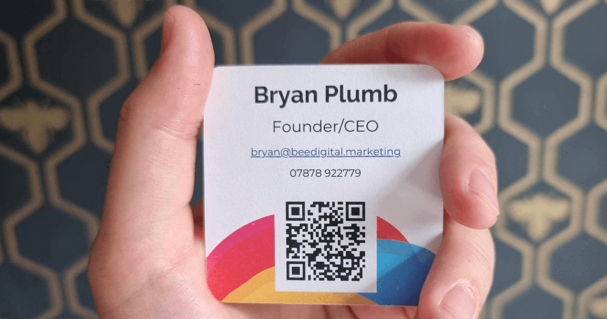 A close up of a hand holding up a square Bee Digital Marketing business card with Bryan's details written on it, and a QR code at the bottom.