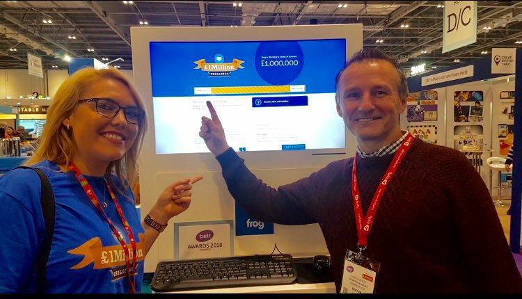 A picture of team members from Frog at the BETT show. A man and a woman stand in front of a computer screen, pointing at the totaliser displayed on it, proud of the money they are saving schools
